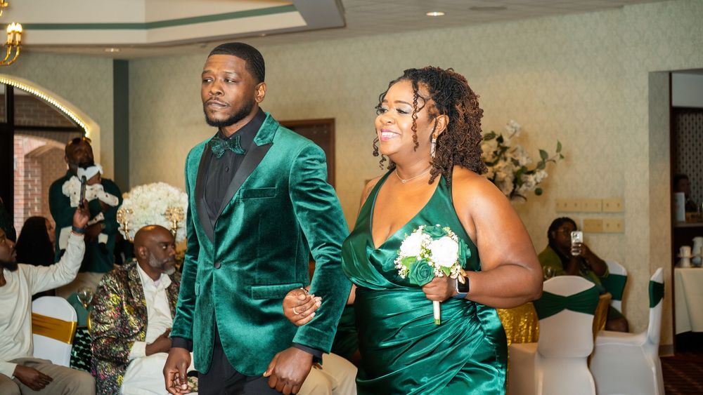 A groomsman in a green velvet suit escorts a bridesmaid in a matching green gown down the aisle at a wedding reception.