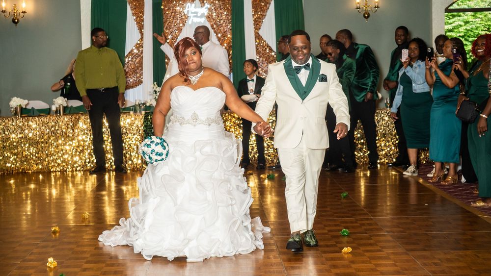 Bride in white ruffled gown and groom in cream tuxedo with green accents walk hand-in-hand at wedding reception.