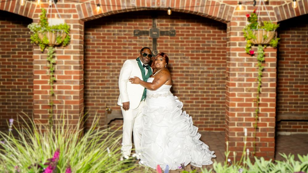 Bride and groom posing in front of brick chapel with cross, surrounded by lush greenery and flowers on their wedding day.
