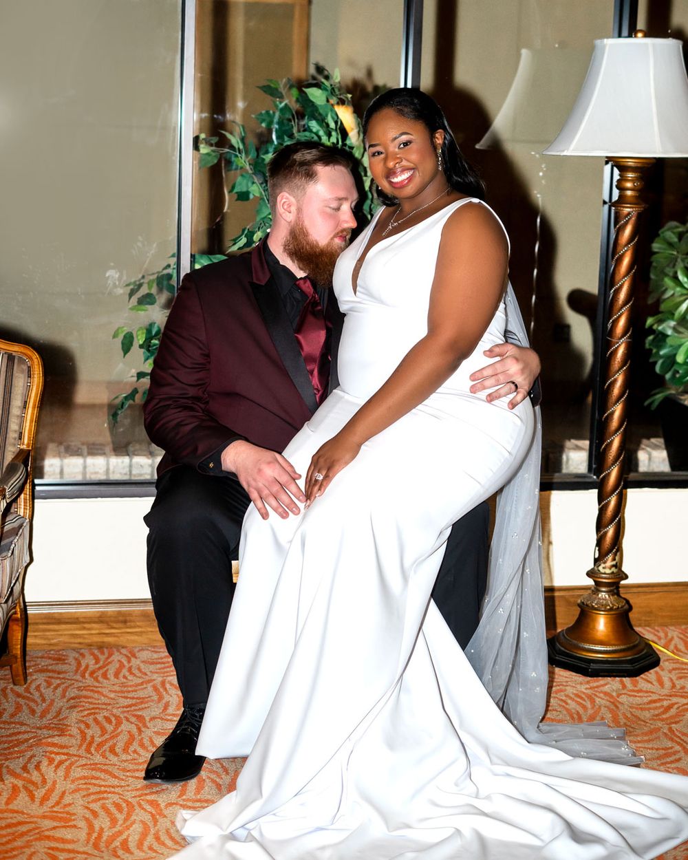 A smiling couple poses together at a formal event, with one in a burgundy suit and one wearing an elegant white gown.