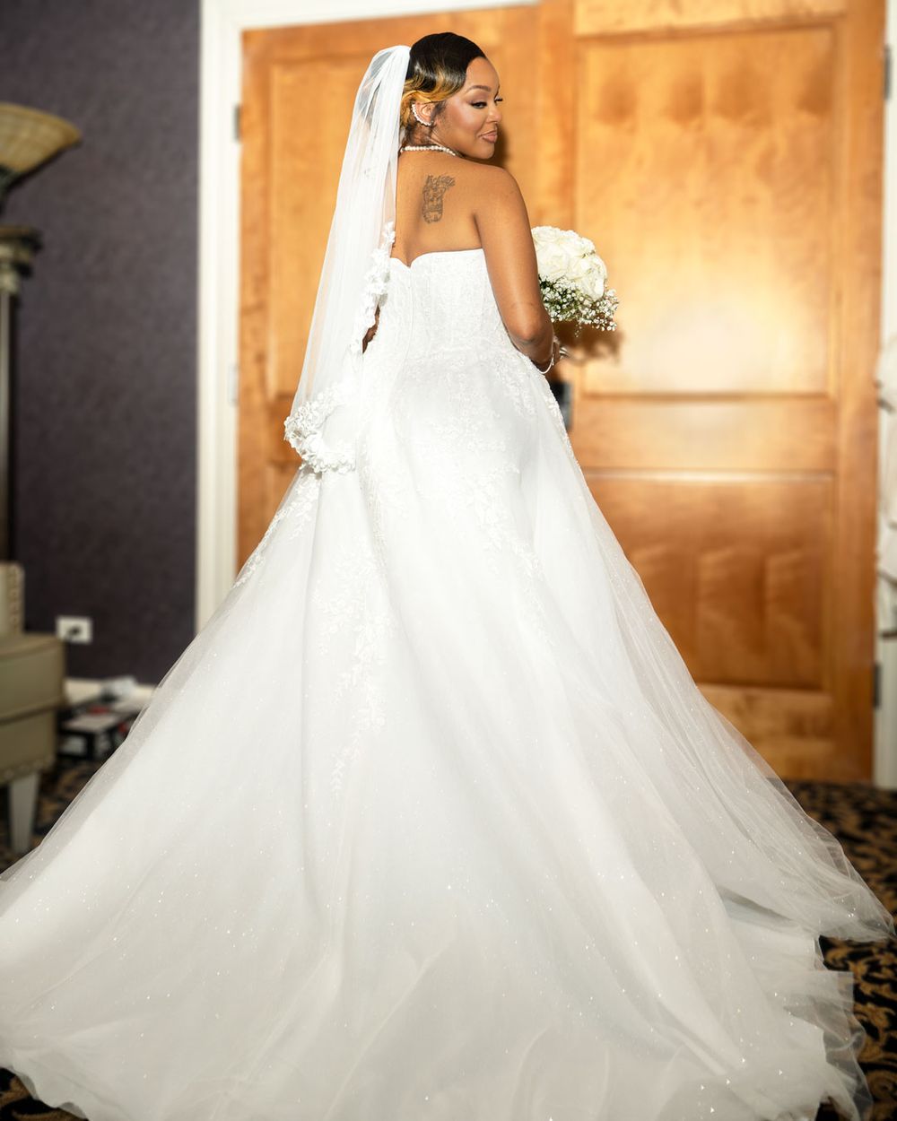 Bride in a strapless white ball gown with lace details, long veil, and floral bouquet, posing with back to camera.