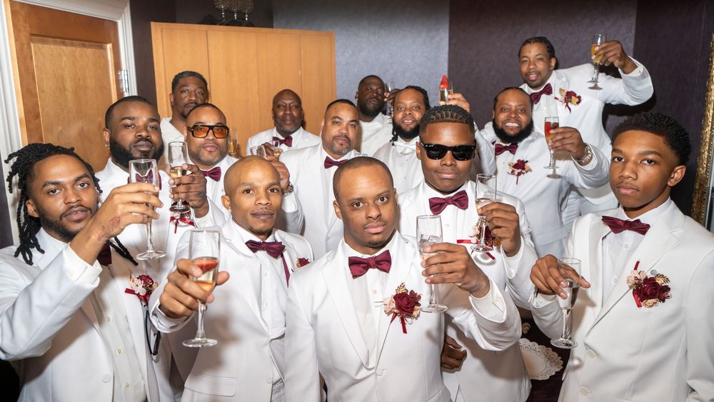 Groomsmen in white tuxedos with burgundy boutonnieres raise champagne glasses in a celebratory toast at a wedding.