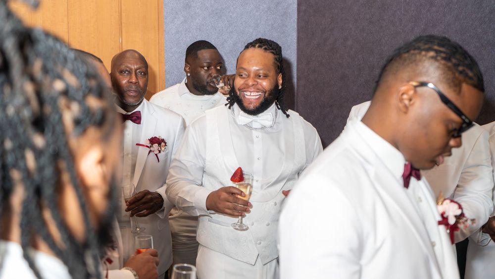 Groomsmen in white suits with burgundy boutonnieres laughing and celebrating at a wedding reception.