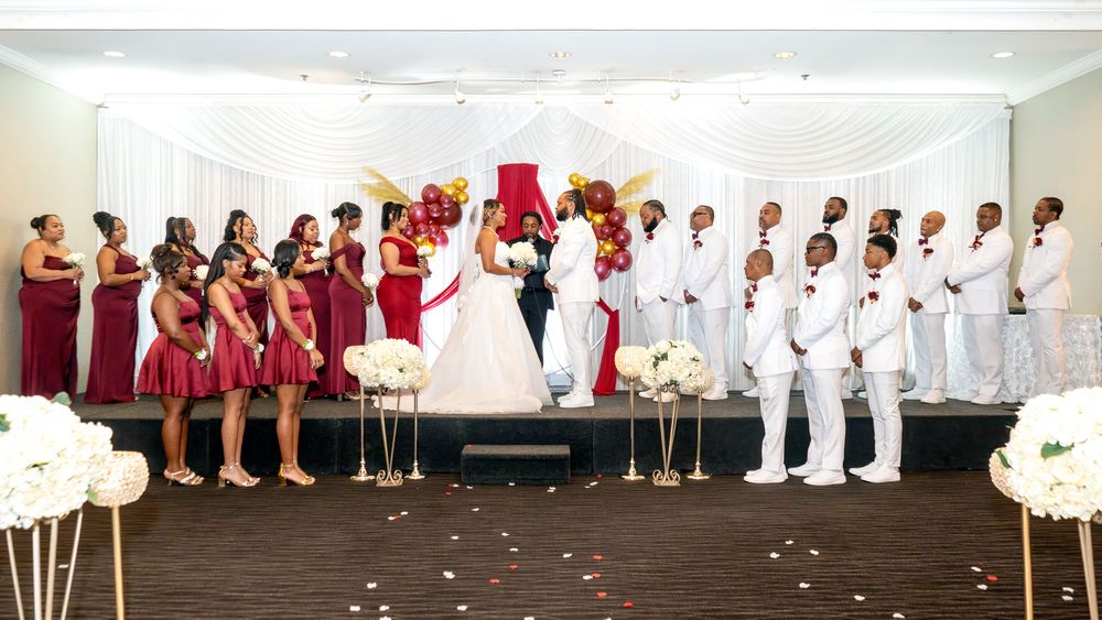 Elegant wedding ceremony with bride, groom, bridesmaids in burgundy dresses, and groomsmen in white suits before draped backdrop.