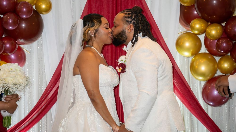 Couple sharing a kiss at wedding altar with red and gold balloon arch decorations in background.