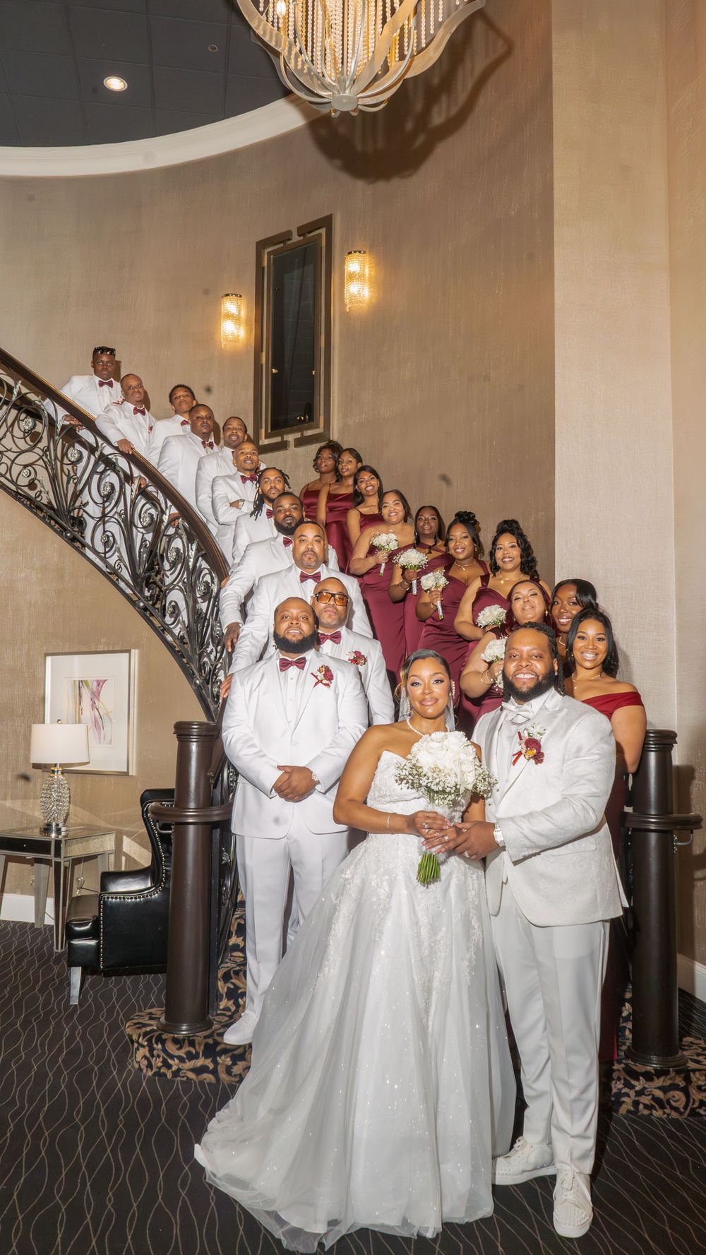 Wedding party poses on elegant staircase, bride in white gown with groom, bridesmaids in burgundy, groomsmen in white suits.