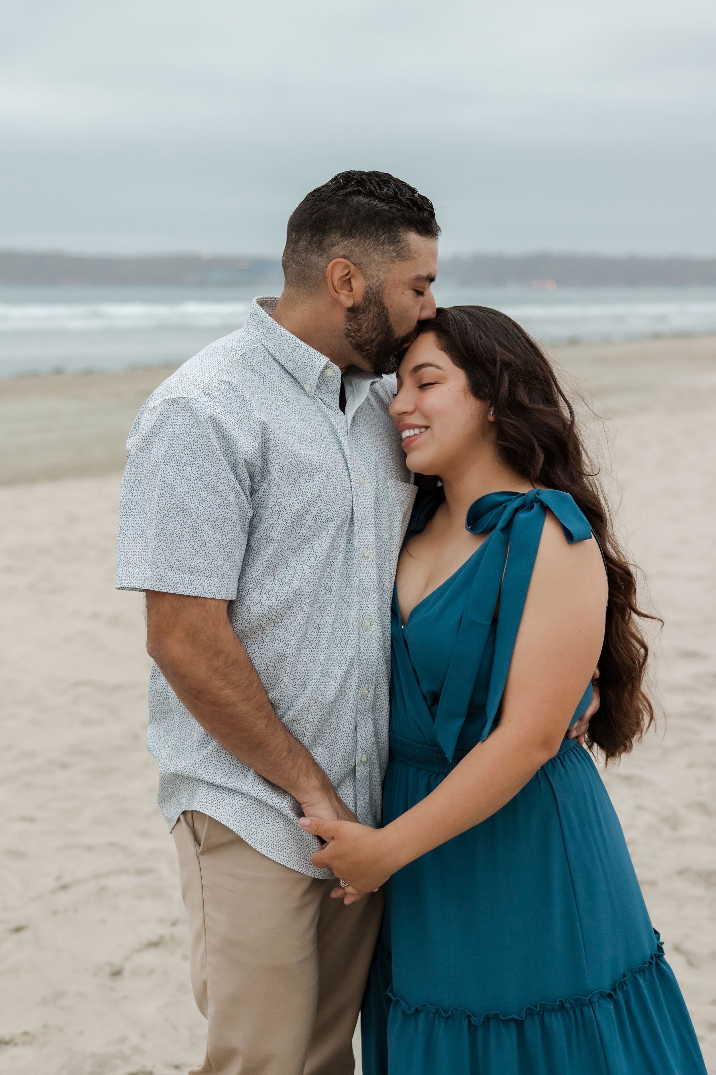 Couple embraces on foggy beach while sharing affectionate moments in casual teal dress and grey shirt.