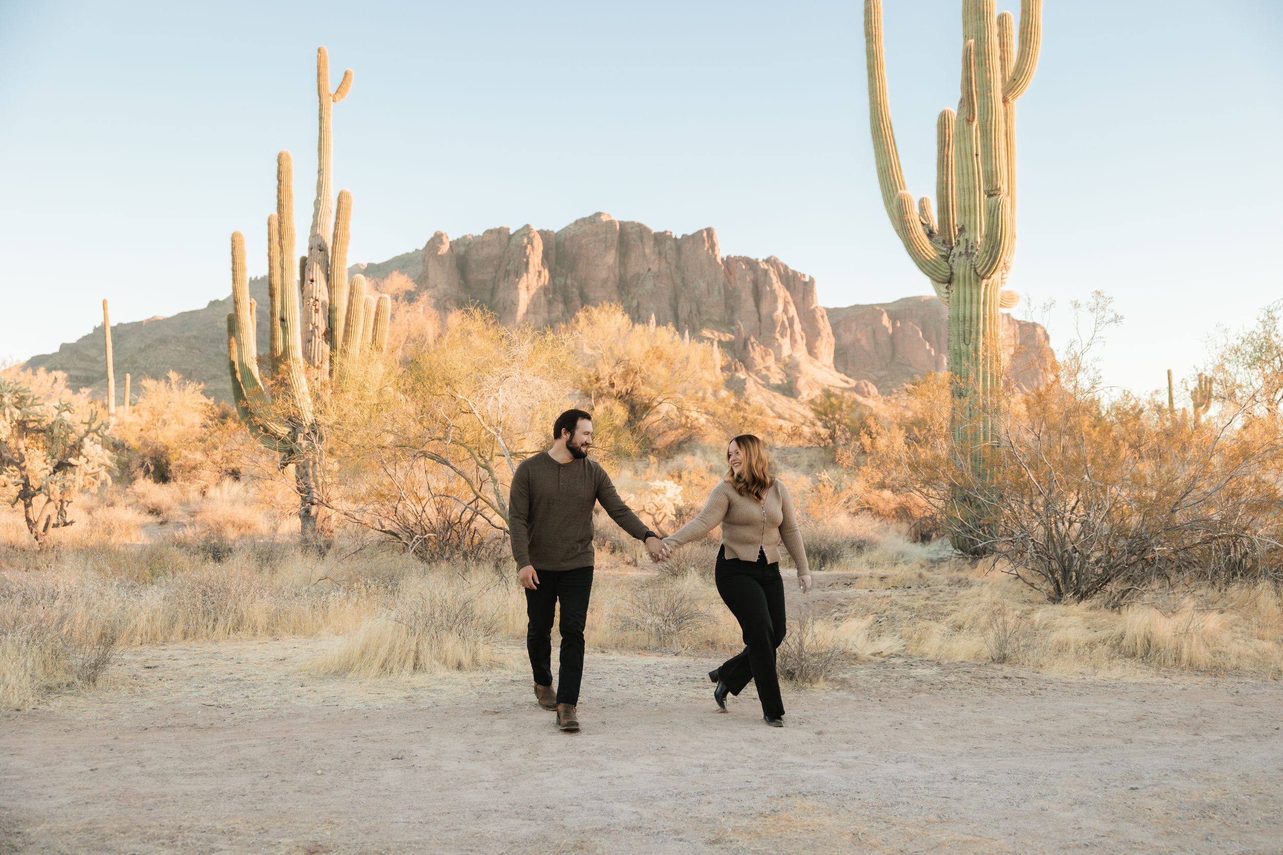 A couple walks through a desert landscape at sunset surrounded by tall saguaro cacti and rocky terrain.
