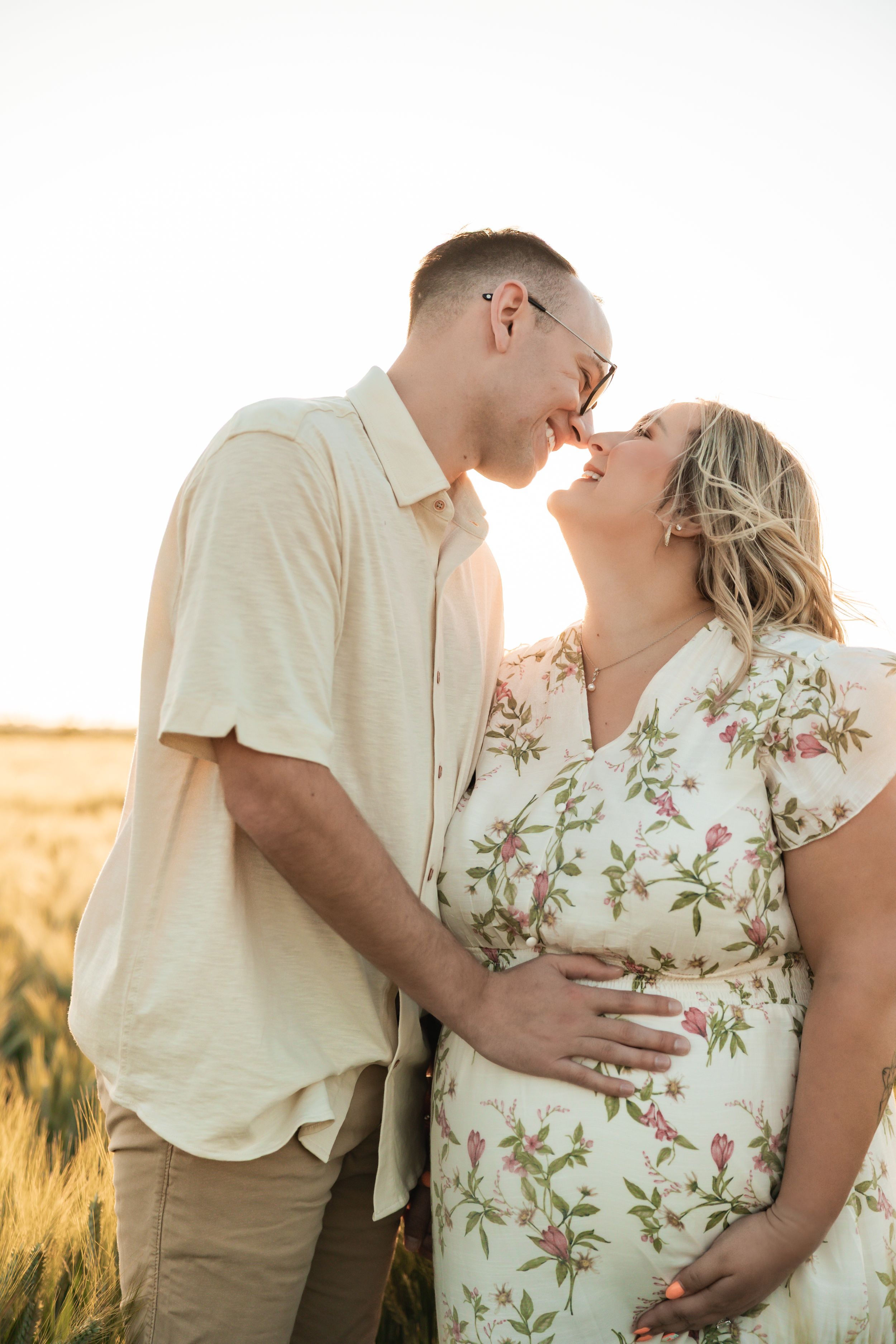 A romantic couple in white and khaki stand close together in a sun-lit golden wheat field during sunset.