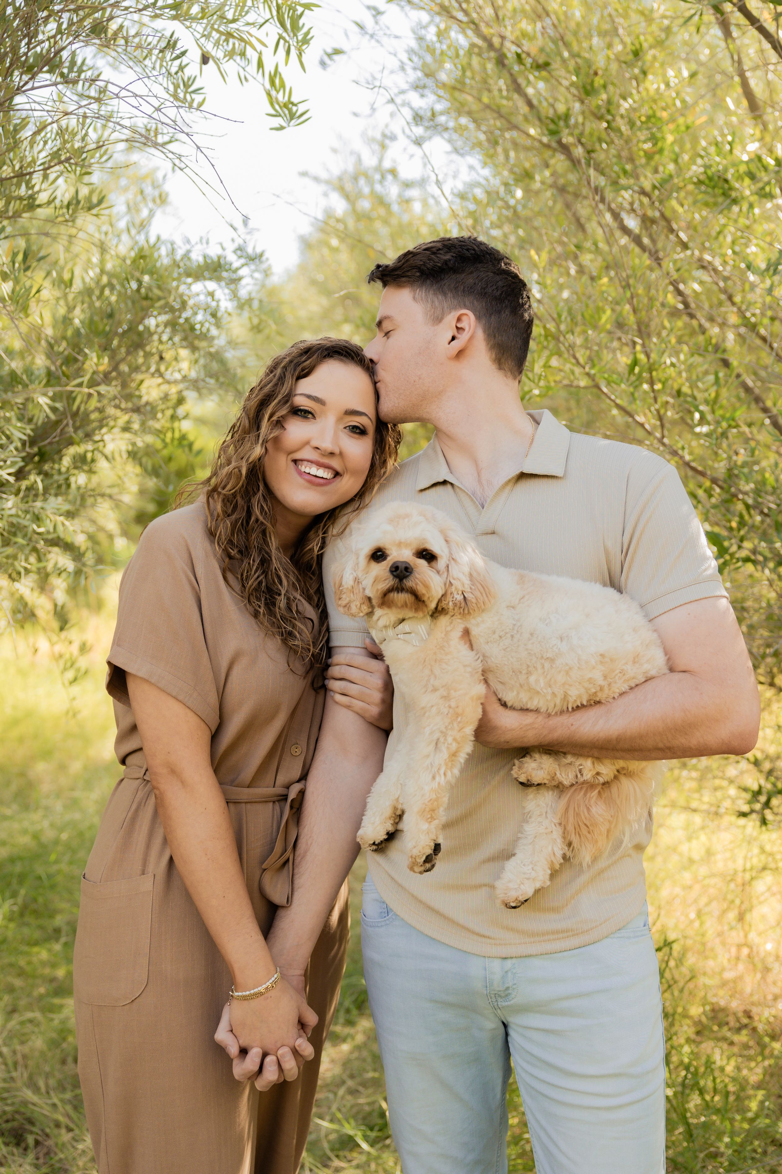Couple enjoying outdoor photoshoot in nature with their small white dog against golden sunlit background.