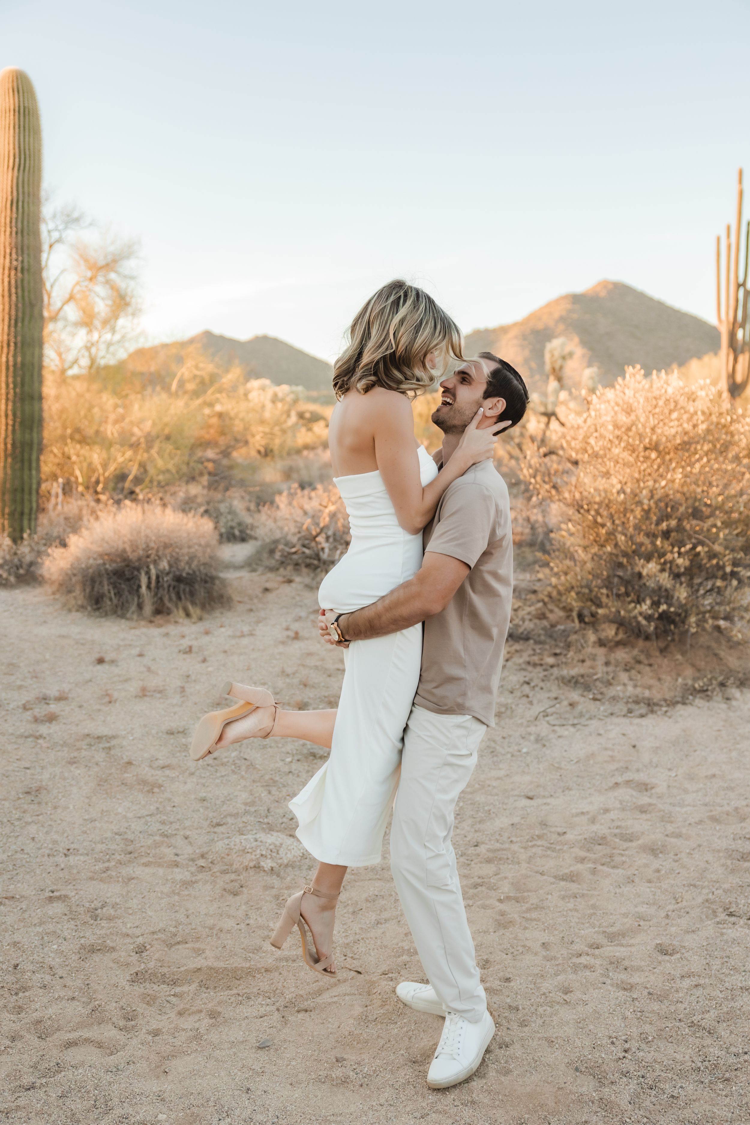 A couple in white outfits share romantic embraces in a desert landscape with saguaro cacti at sunset.