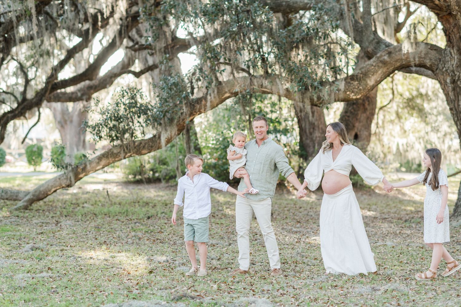 A joyful family walks hand-in-hand under sprawling oak trees draped with Spanish moss outdoors.