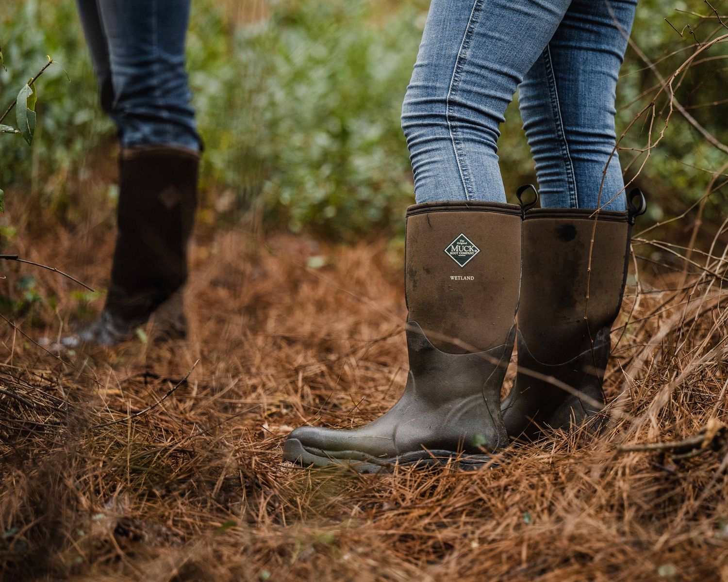 Person wearing blue jeans and brown boots standing on forest ground covered in fallen leaves.