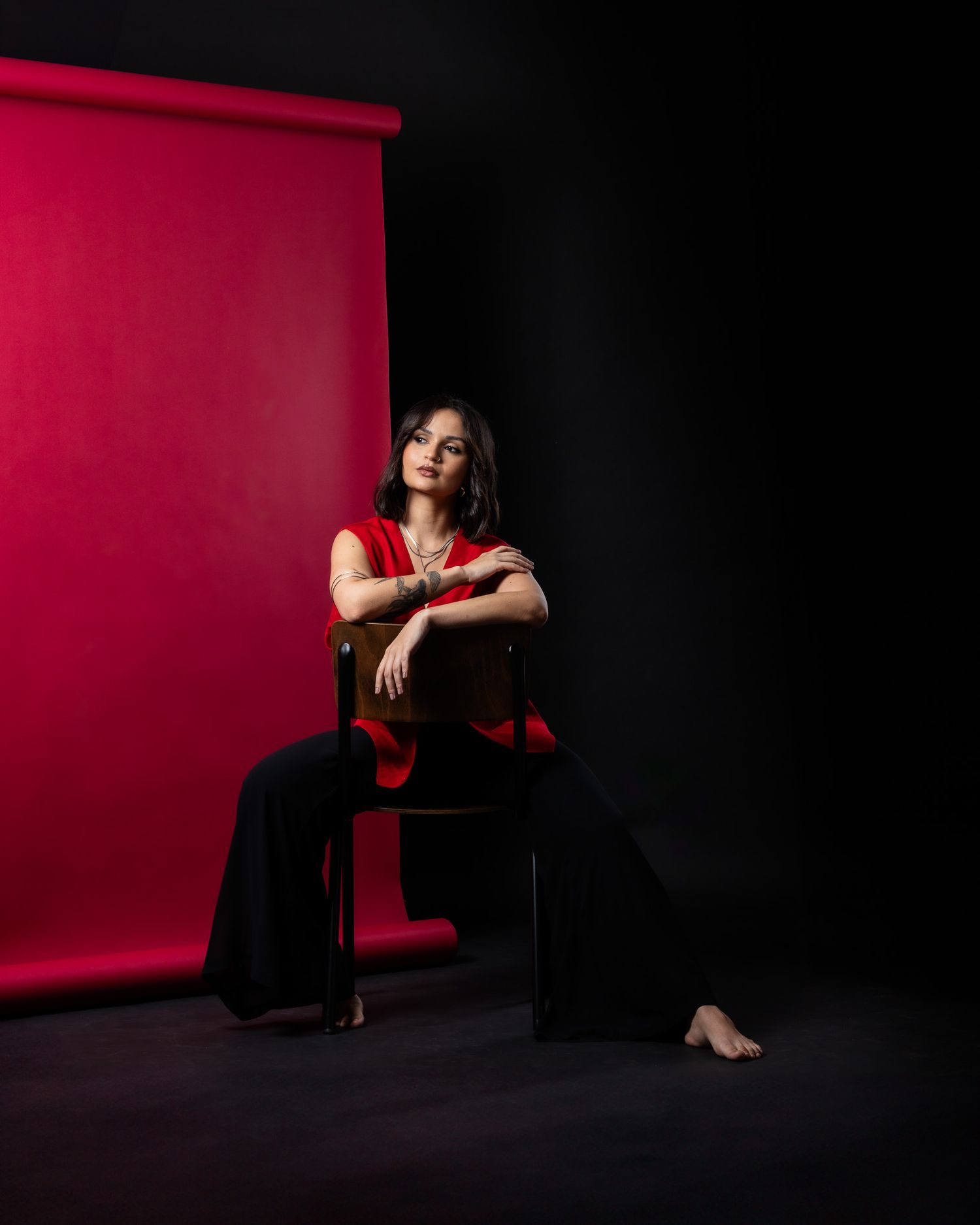 Woman in red top and black pants poses on chair against red backdrop.