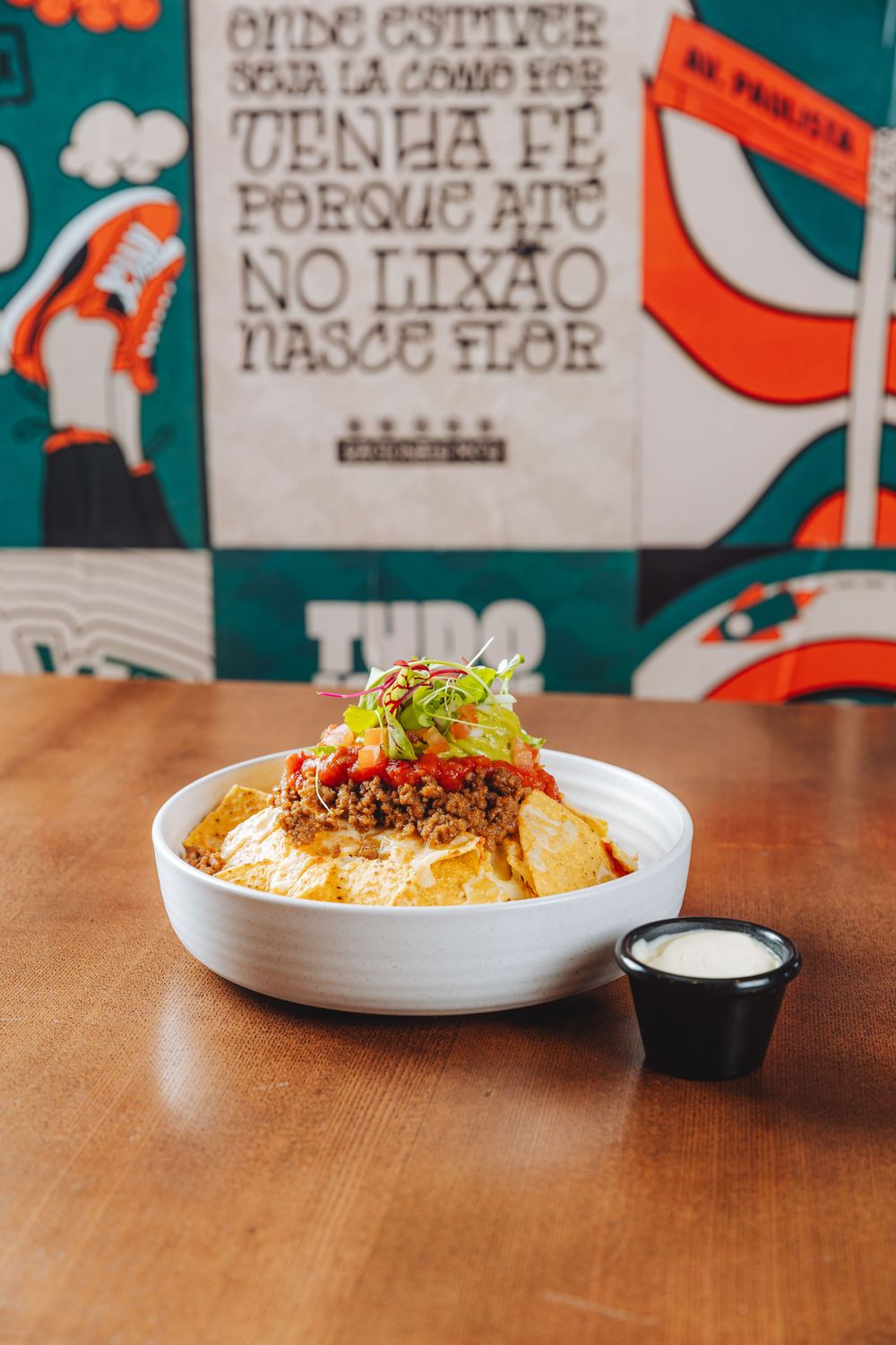 A bowl of loaded nachos topped with meat, cheese sauce, and fresh garnishes served with a side of dip on a wooden table.