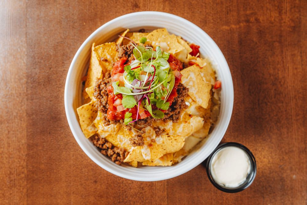 A bowl of loaded nachos topped with melted cheese, ground beef, diced tomatoes, red onions and cilantro, served with sour cream.