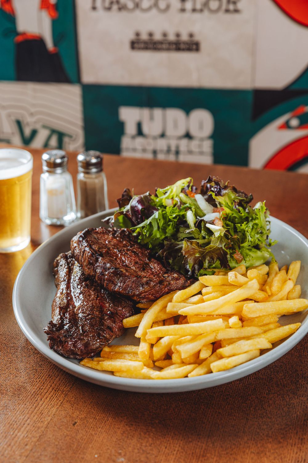 Grilled steak served with crispy french fries and fresh green salad on a gray plate with a beer in the background.