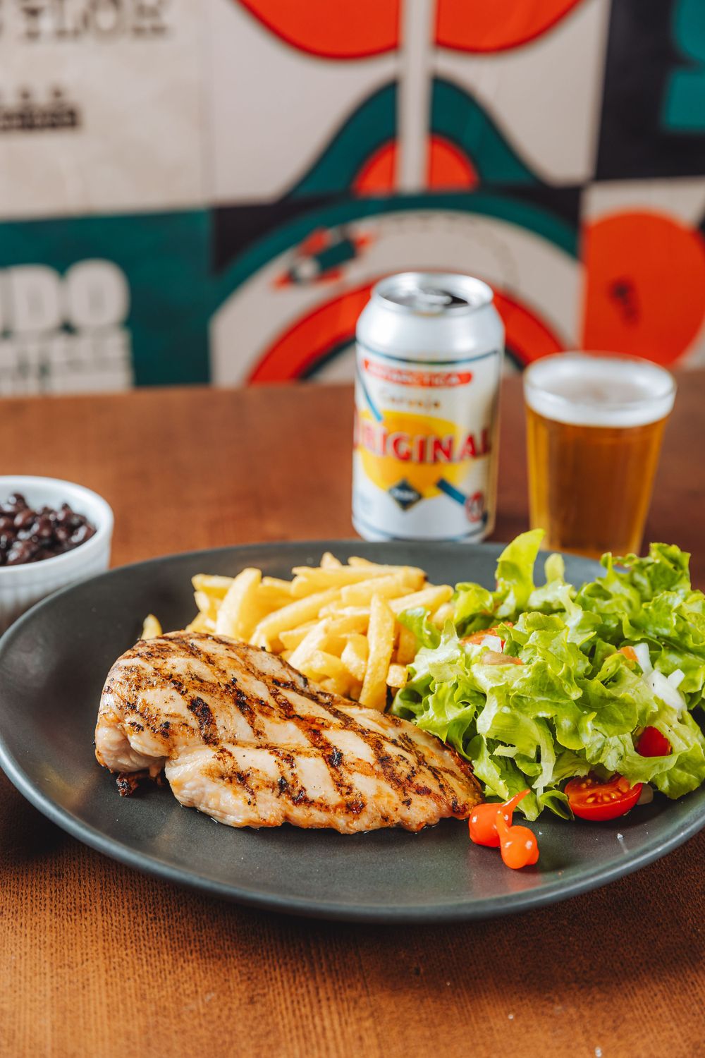 Grilled chicken breast with french fries and garden salad served alongside a cold beer and canned beverage.