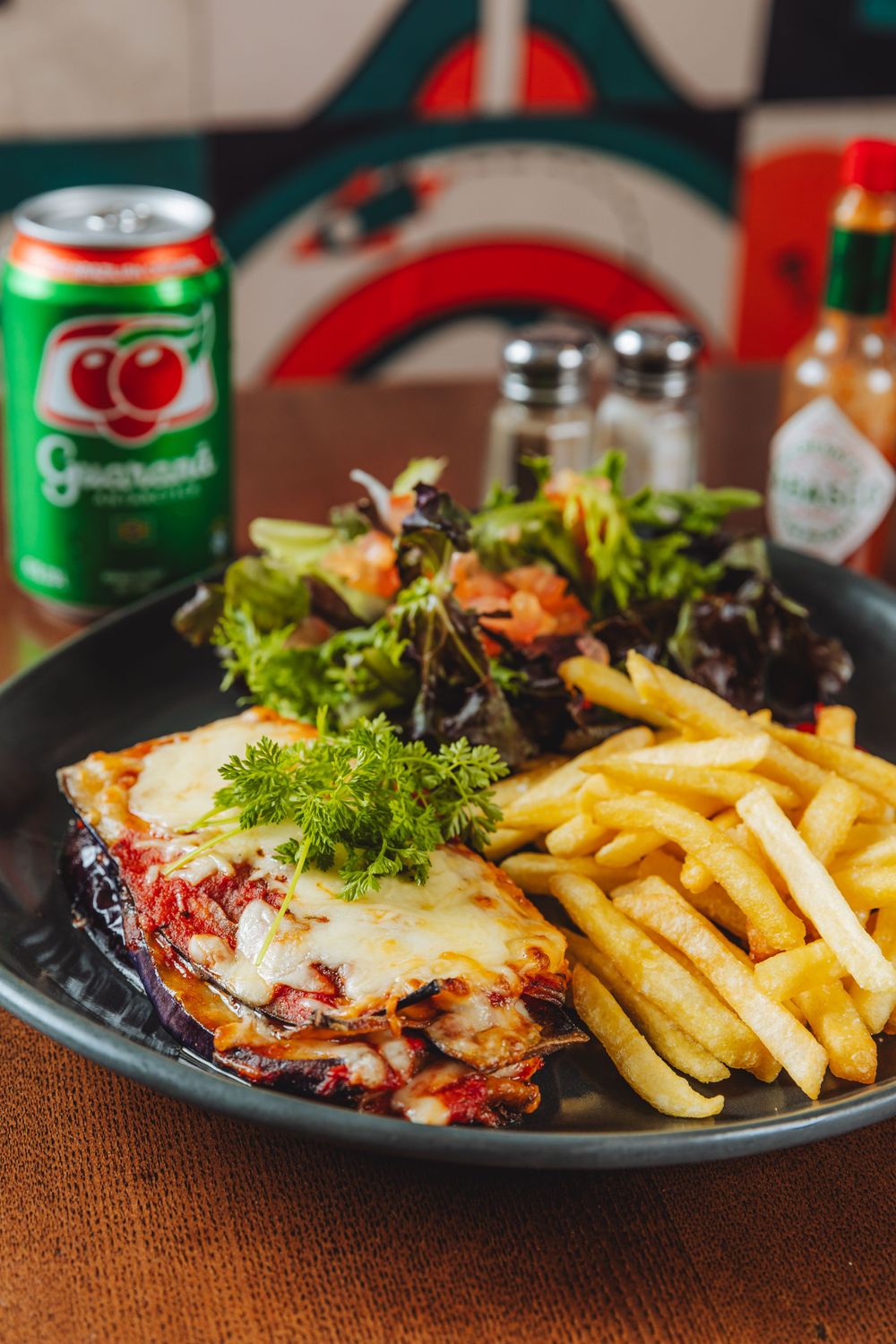 A plate with melted cheese casserole served with crispy french fries, fresh salad and a green soda can in background.