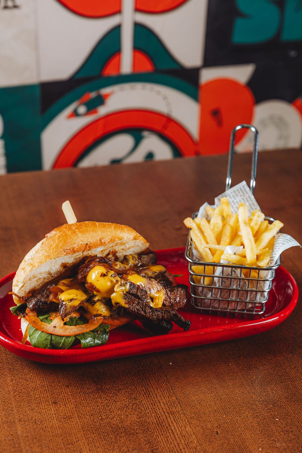 Juicy cheeseburger with melted cheese and french fries served in a wire basket on a red plate against patterned wall decor.