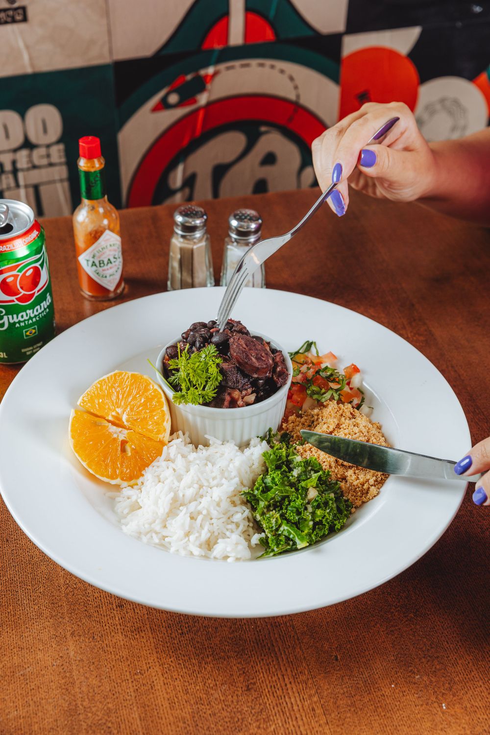 A Brazilian feijoada plate with rice, black beans, orange slice, and leafy greens served alongside Guarana soda.