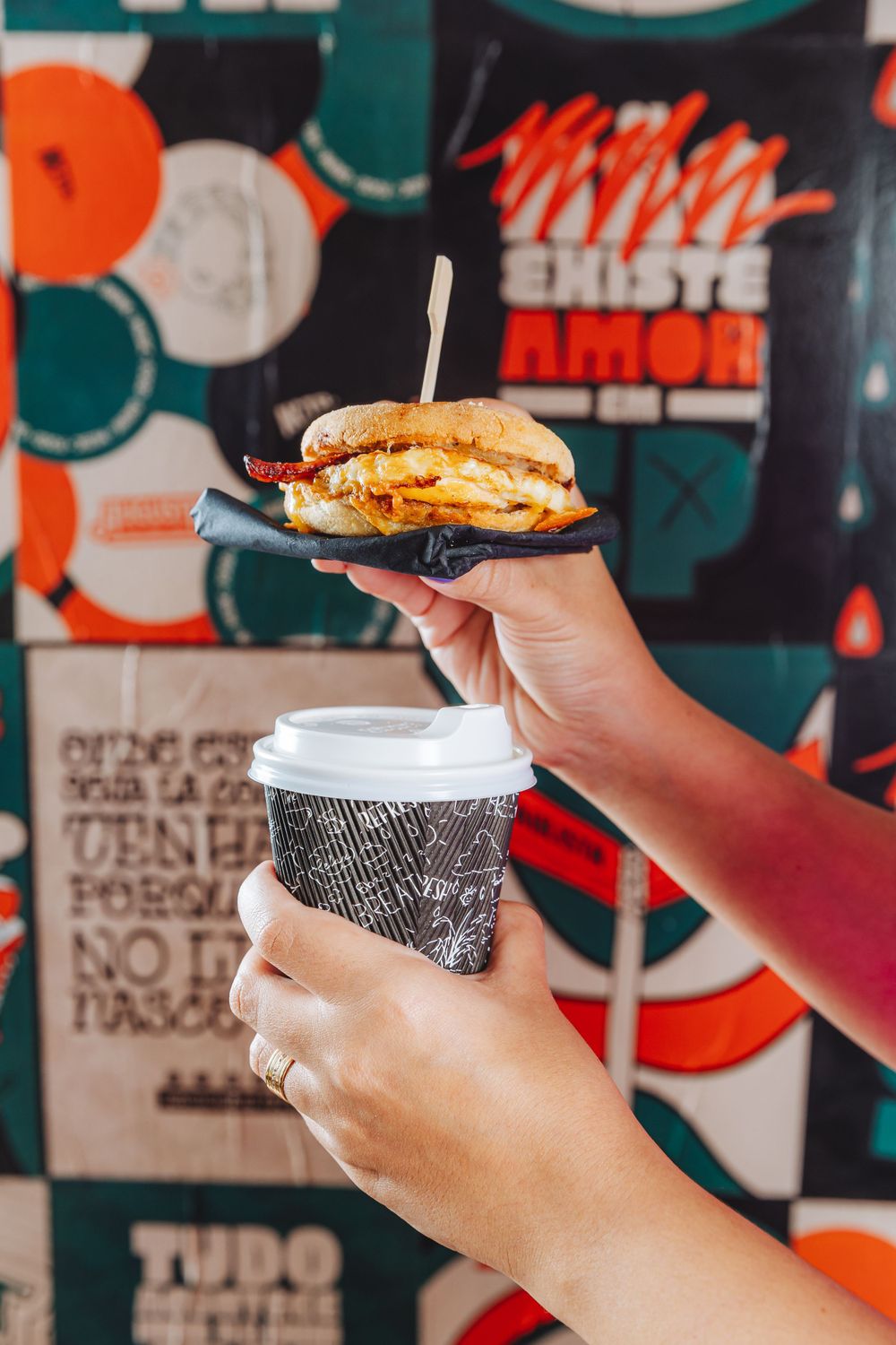 Hand holding a burger and coffee cup against colorful geometric wall art background in casual dining setting.