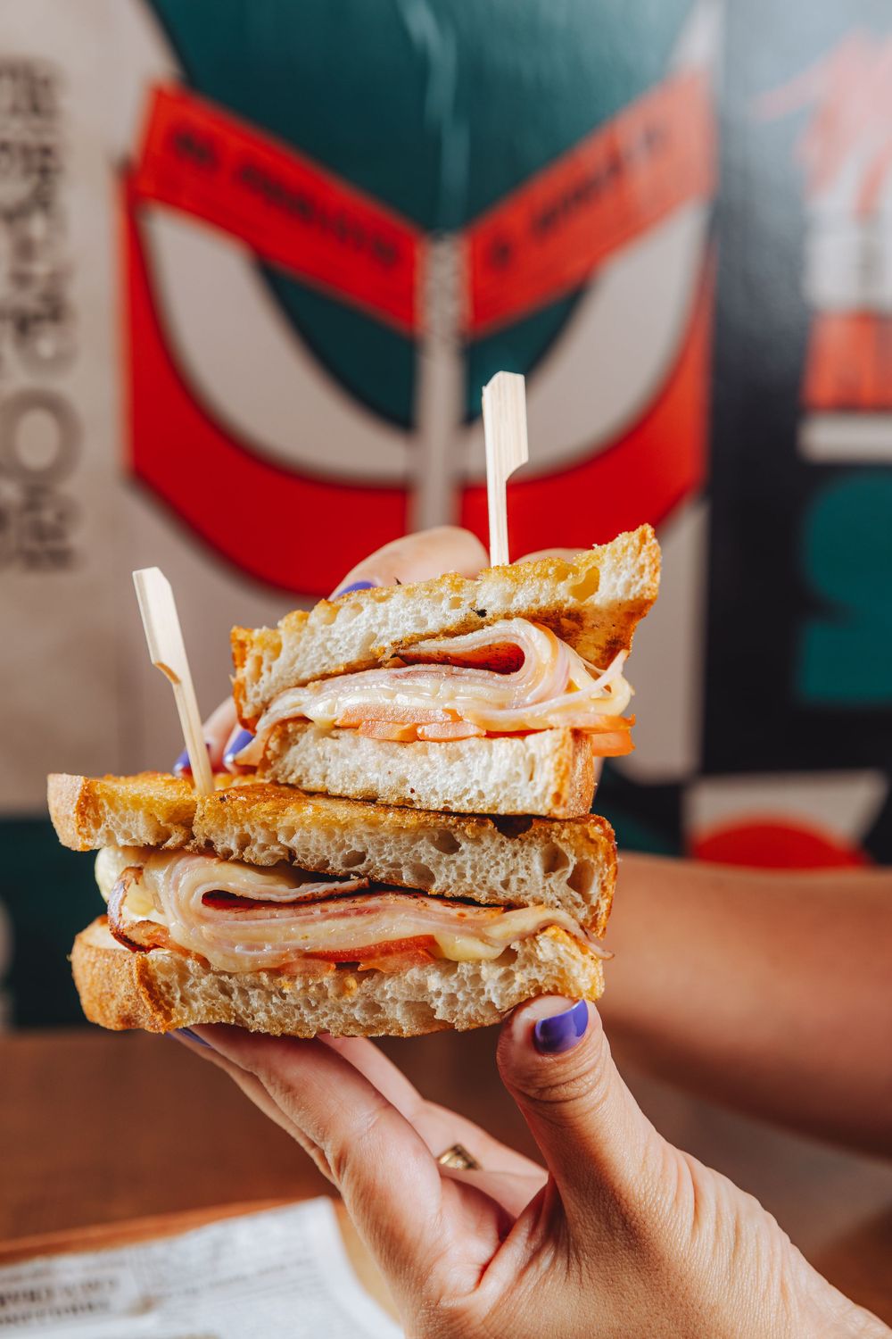 Layered club sandwich with meat and vegetables held up against a colorful wall background.