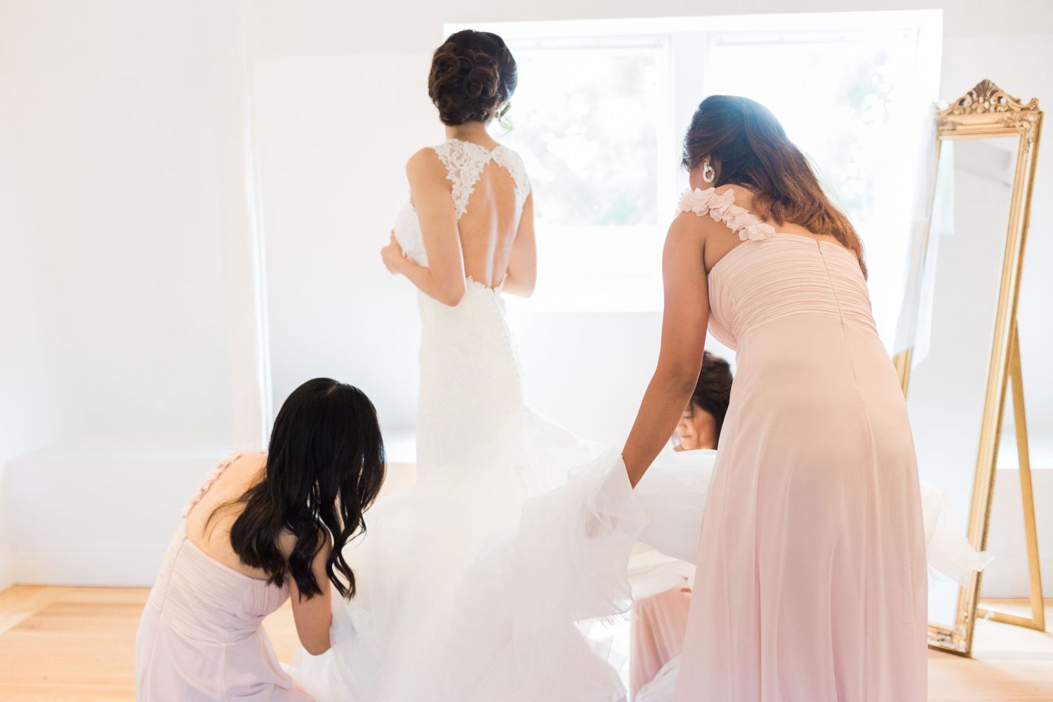 Bridesmaids helping adjust a wedding dress in a bright white room with a gold mirror.