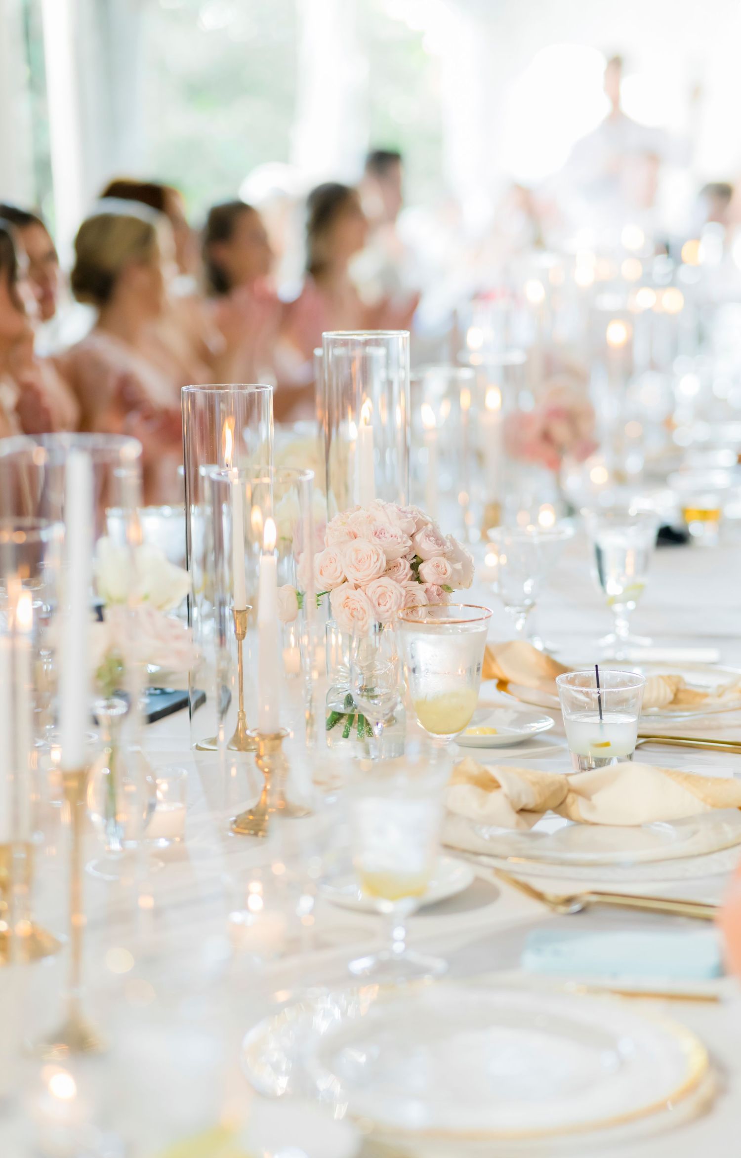 Elegant wedding reception table setting with soft pink floral centerpieces, crystal glasses and candlelight on white linens.