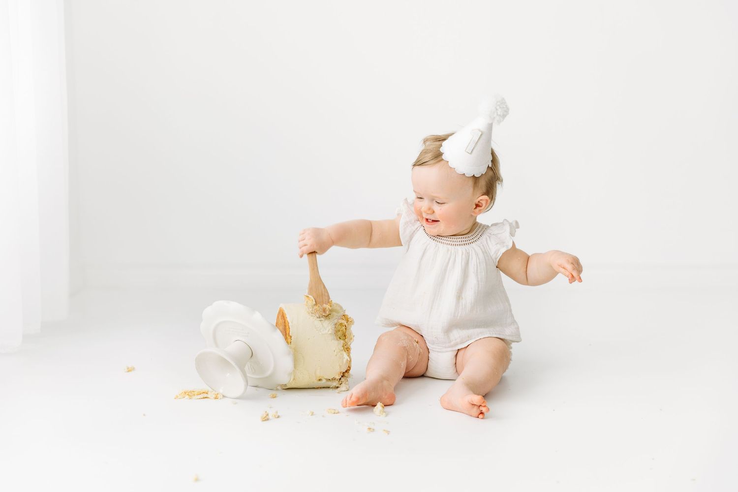 First Birthday portrait, baby eating cake with wooden spoon, wearing birthday hat