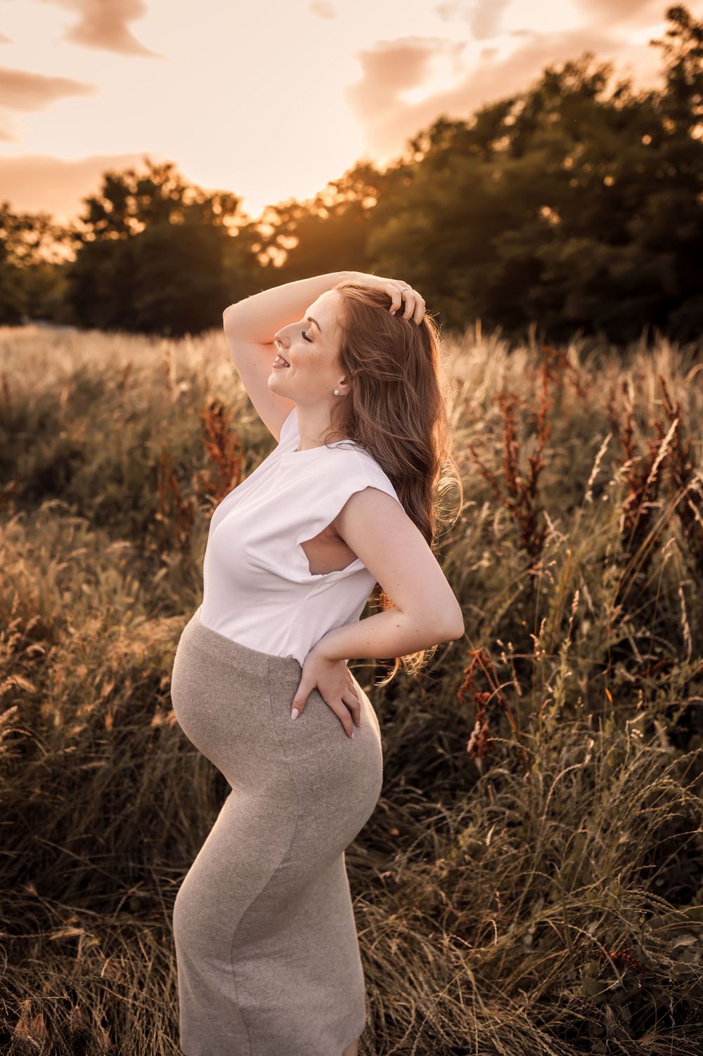 An expectant mother poses in a field at sunset wearing a white top and gray dress with warm golden lighting.