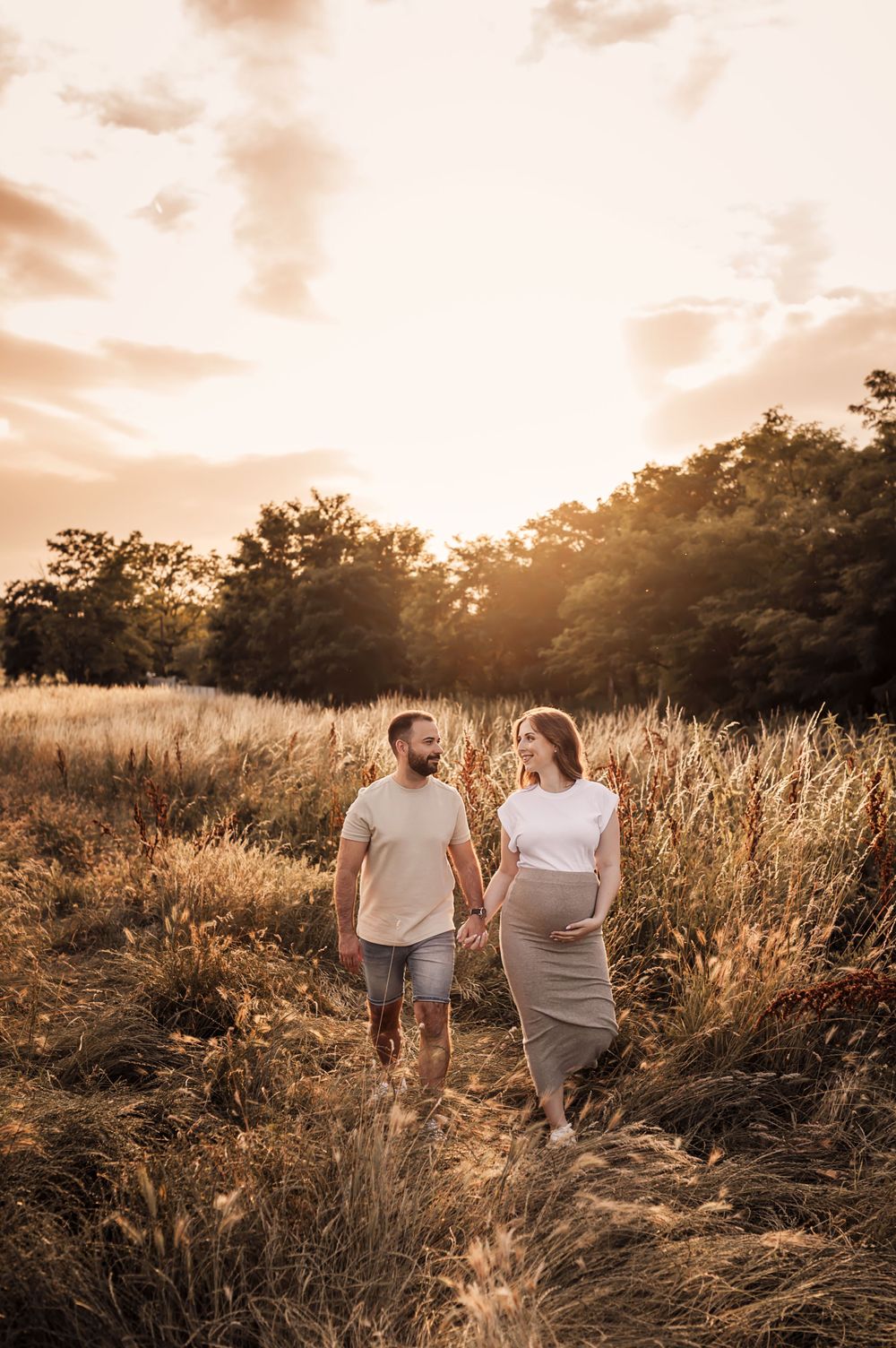 A couple stands together in a golden wheat field at sunset, backlit by warm sunlight filtering through trees.
