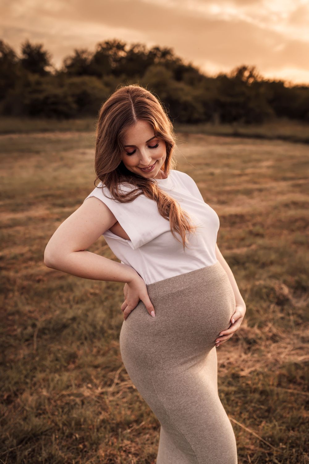 A maternity portrait at sunset with a woman in a white top and beige skirt standing in a grassy field.