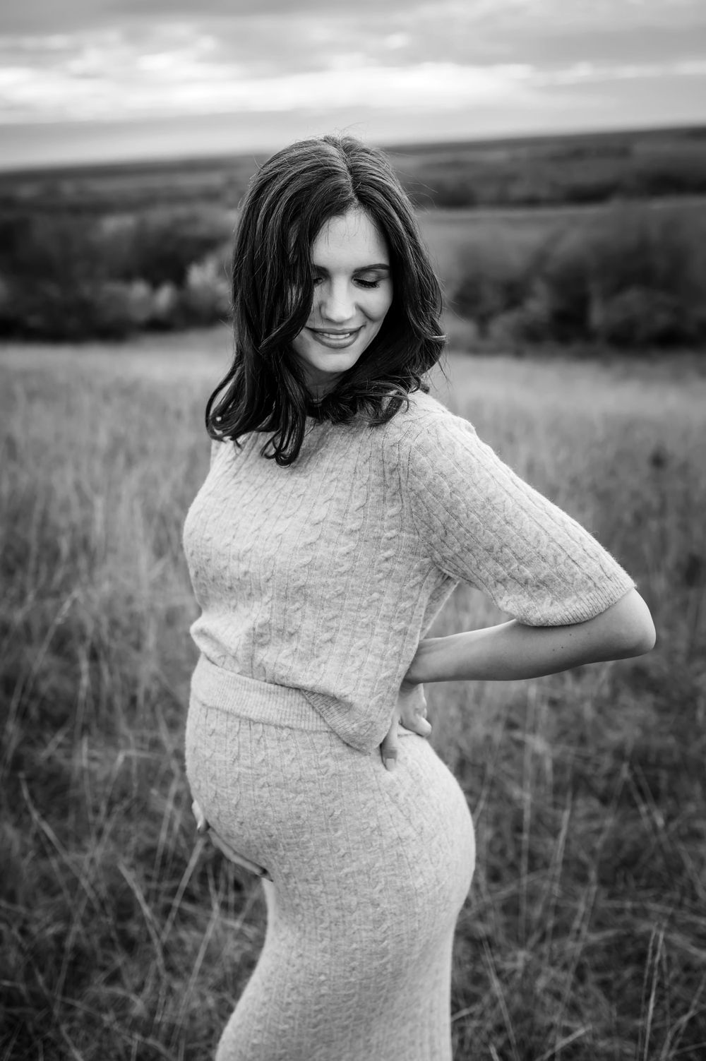 A pregnant woman in a white sweater dress stands in a field on an overcast day, smiling with long dark hair.