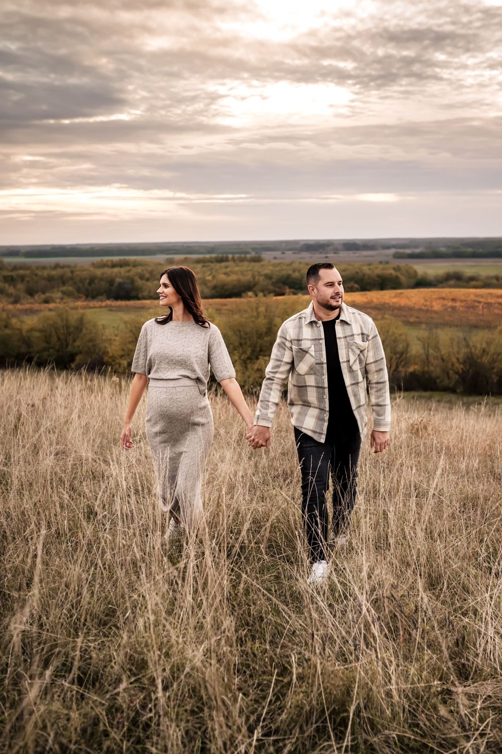 A couple holds hands while walking through a golden meadow at sunset with cloudy skies overhead.