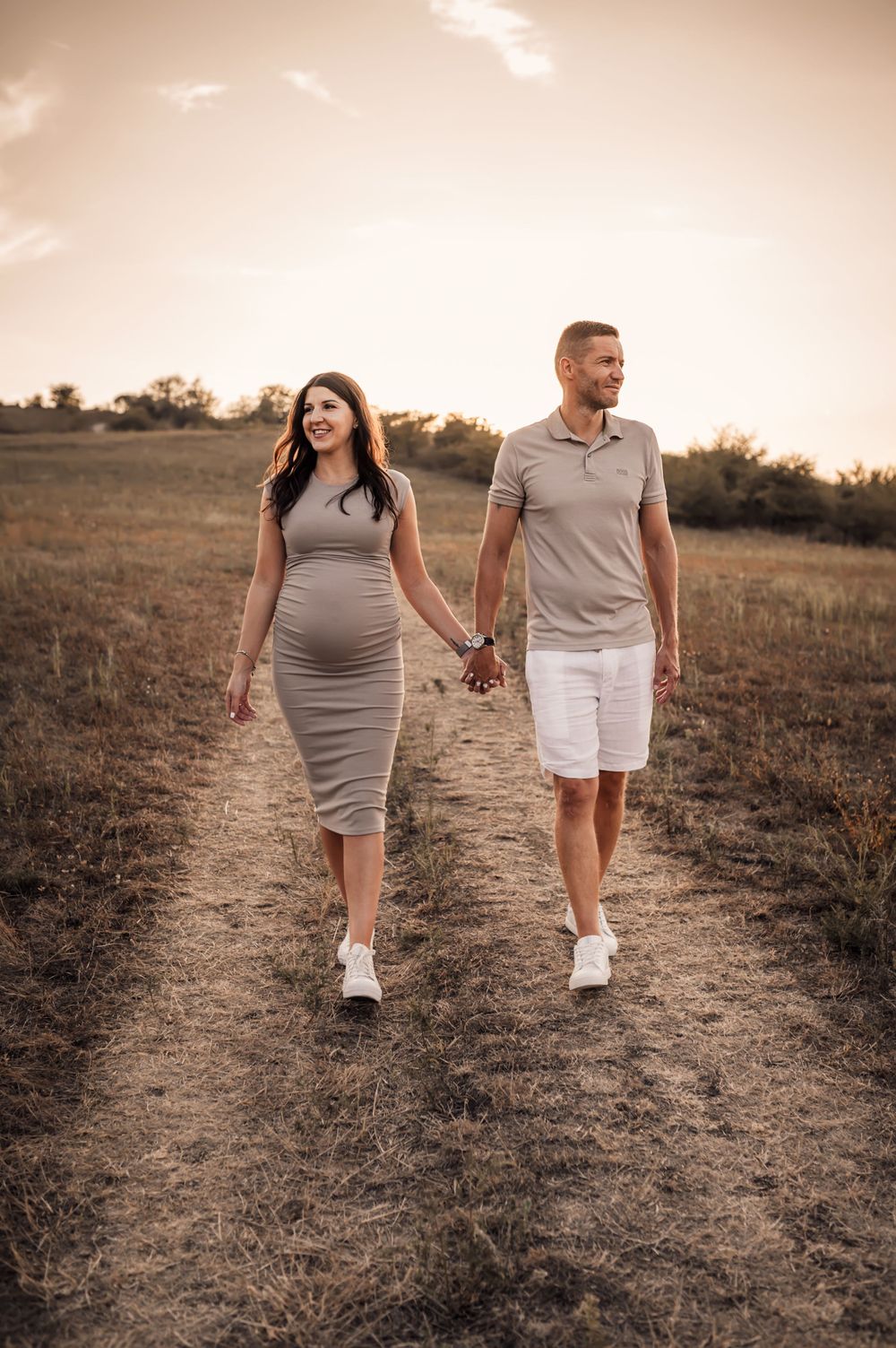 A couple walks hand in hand along a dirt path during sunset in a rustic outdoor setting.