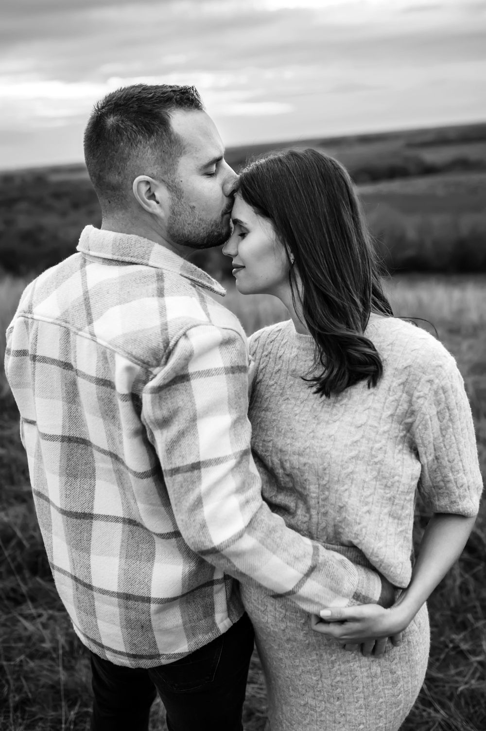 A romantic black and white portrait of a couple embracing in a sweater and plaid shirt against a moody outdoor landscape.
