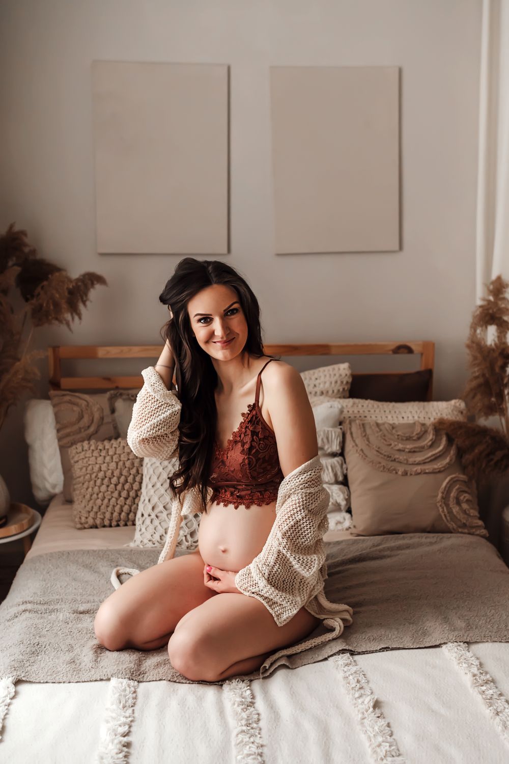 A woman in red lingerie and cozy cardigan poses on a bed with neutral decor and pampas grass accents.
