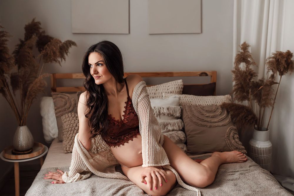 A woman in burgundy lingerie reclines on a bed with neutral decor and pampas grass arrangements in the background.