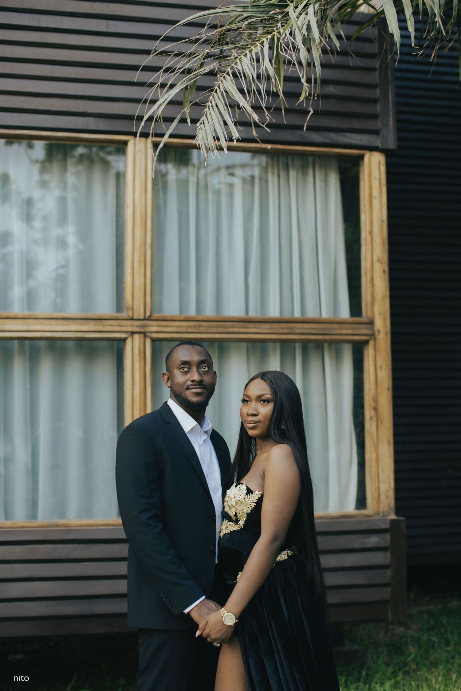Elegant couple poses against wooden window frame in formal evening attire.