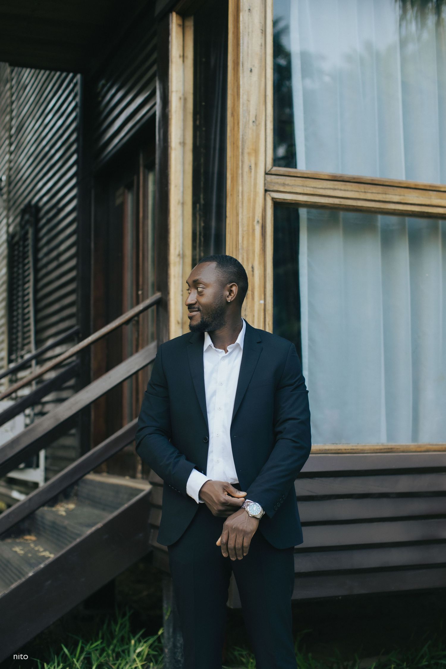 Professional wearing a dark suit stands by a modern building with wooden accents and glass windows.