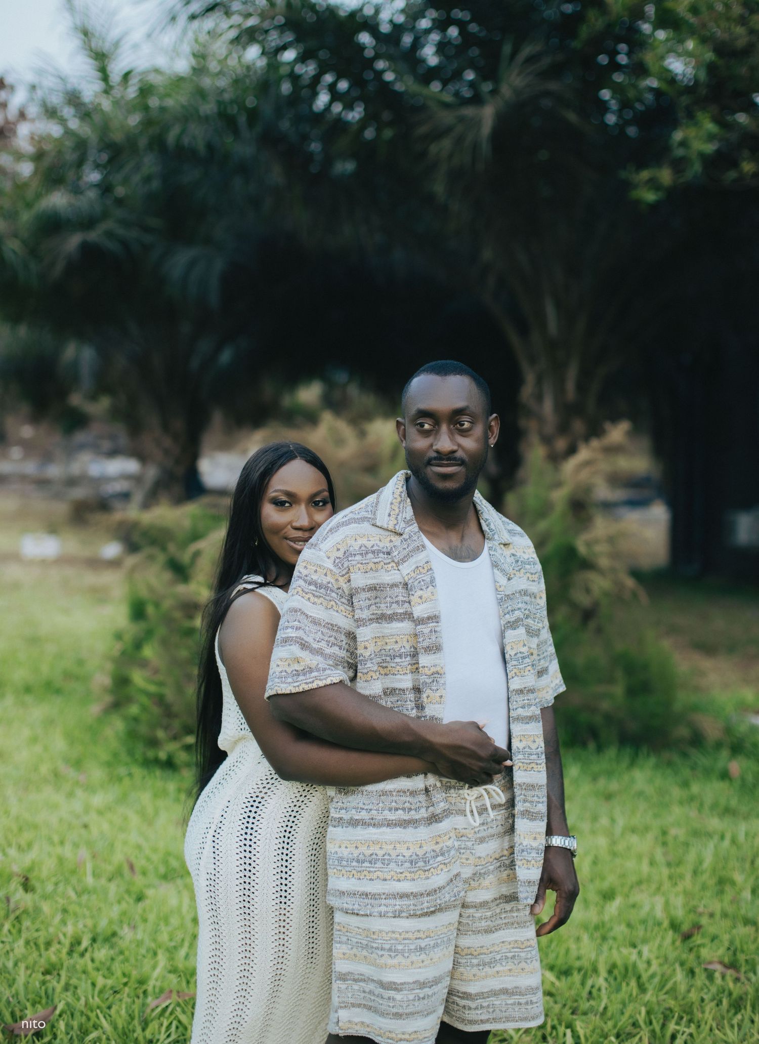 A couple poses together outdoors wearing matching cream and white patterned casual outfits.