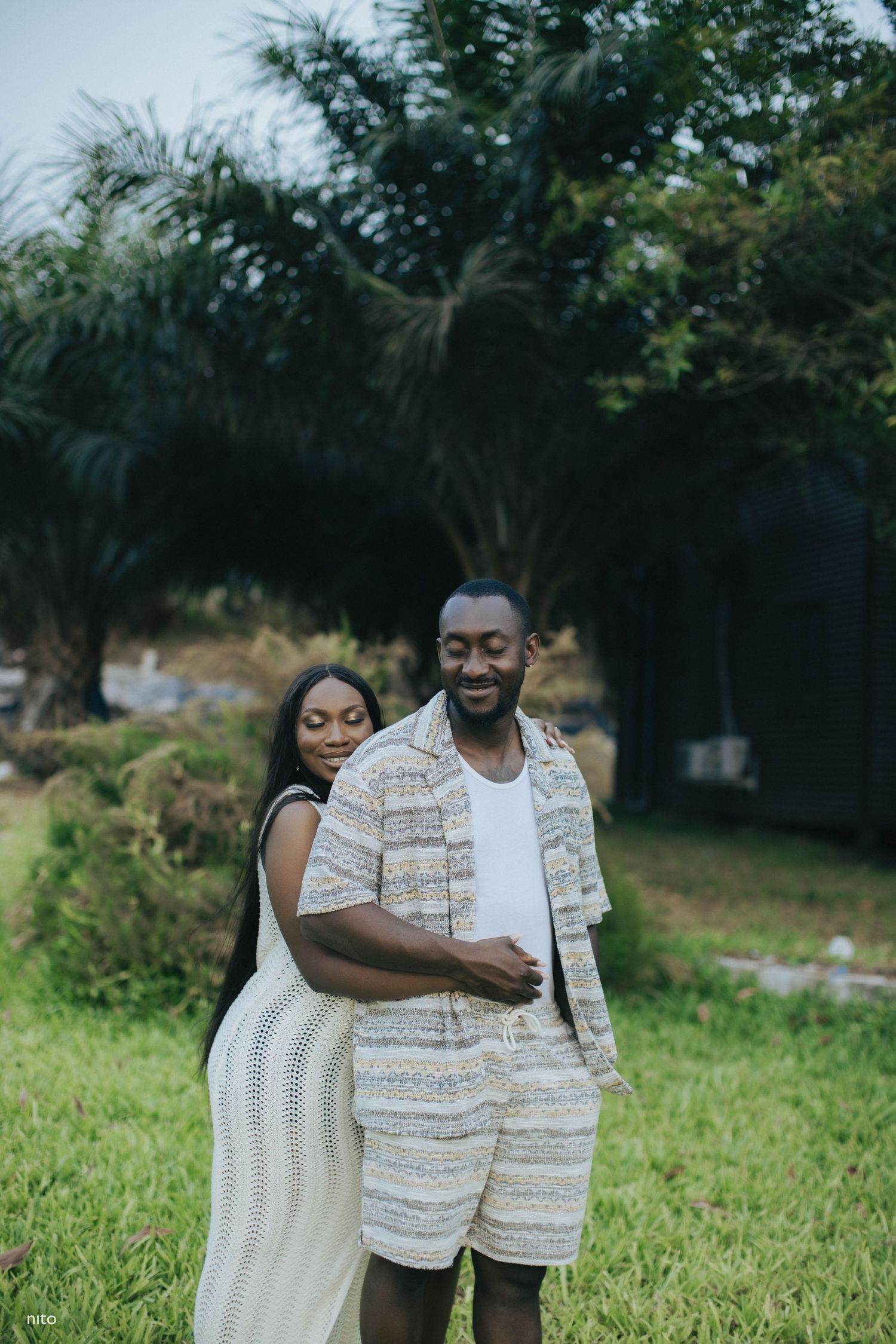 A couple embraces in casual cream-colored outfits while standing in a lush garden setting.