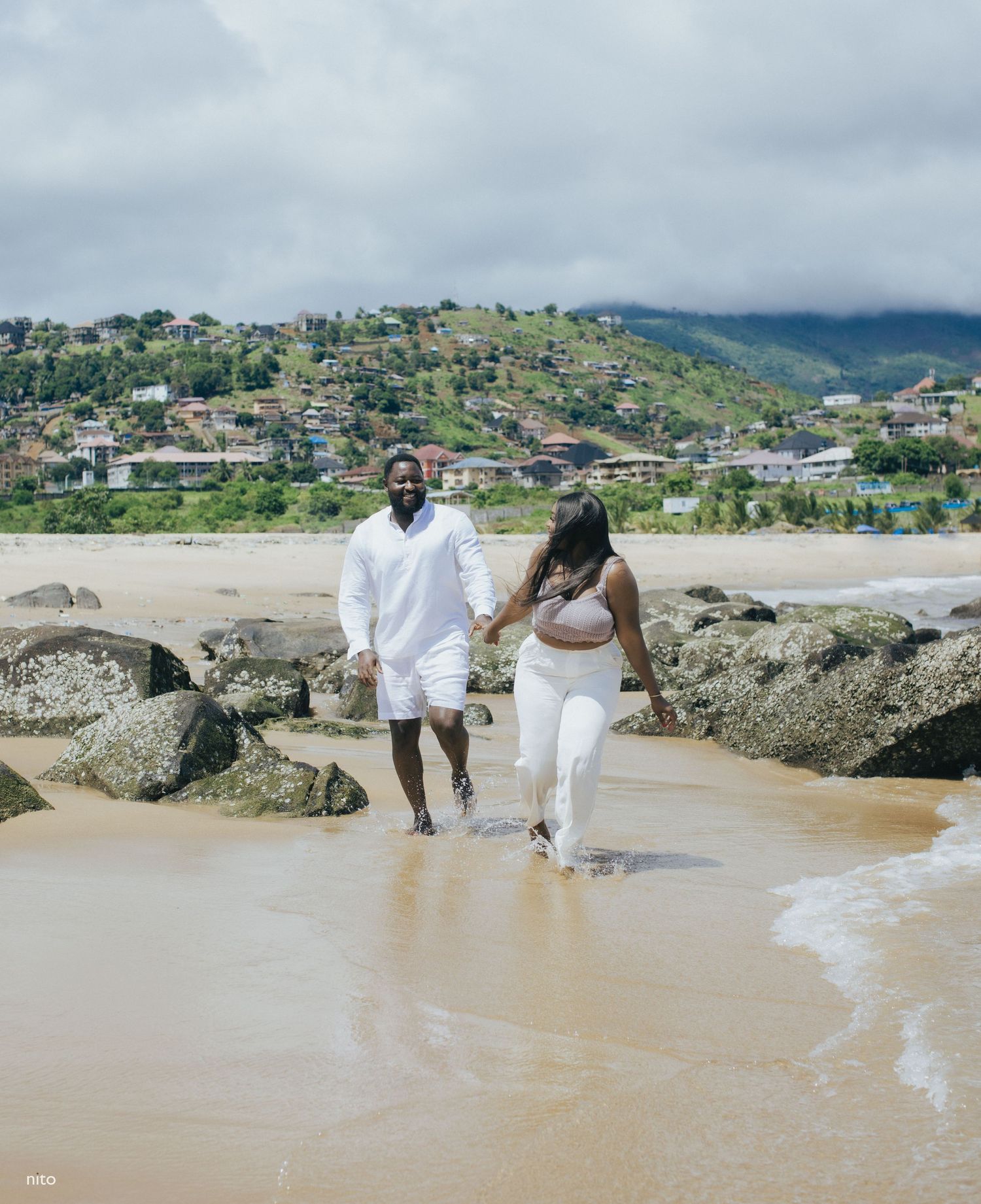 A couple walks along a beach with green hills and coastal homes in the background.