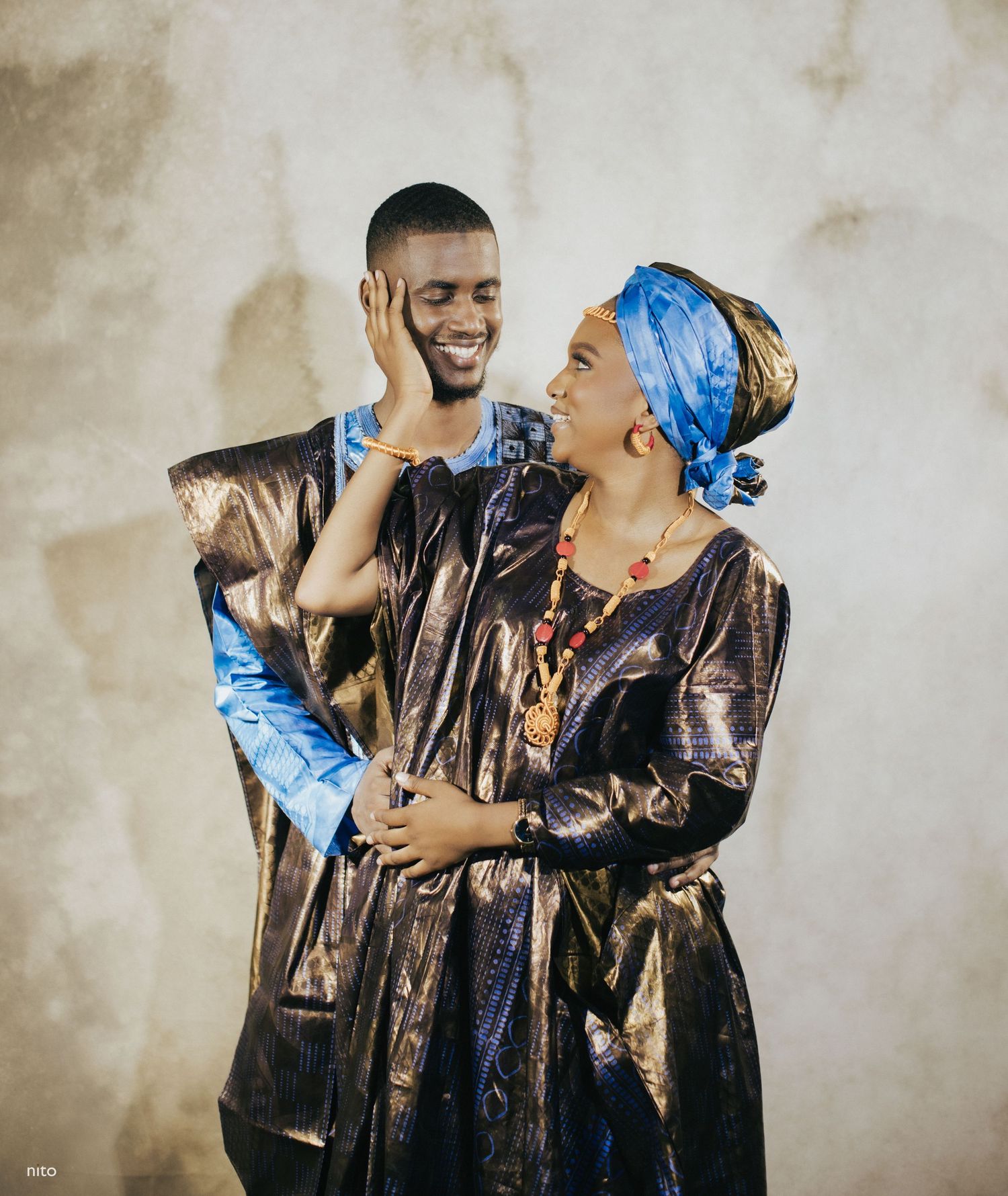 A couple in matching metallic African garments share a playful moment against a neutral wall.