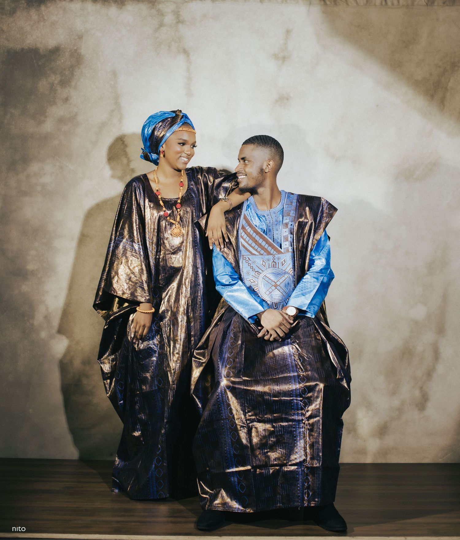 A couple in traditional African clothing sits elegantly against a neutral studio backdrop.