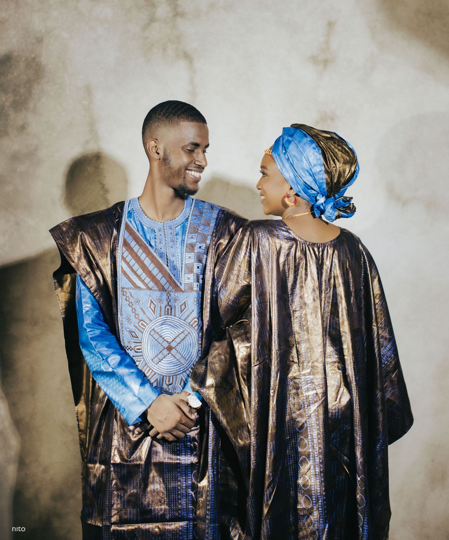 A couple in coordinating blue and bronze African formal wear pose against a textured wall.