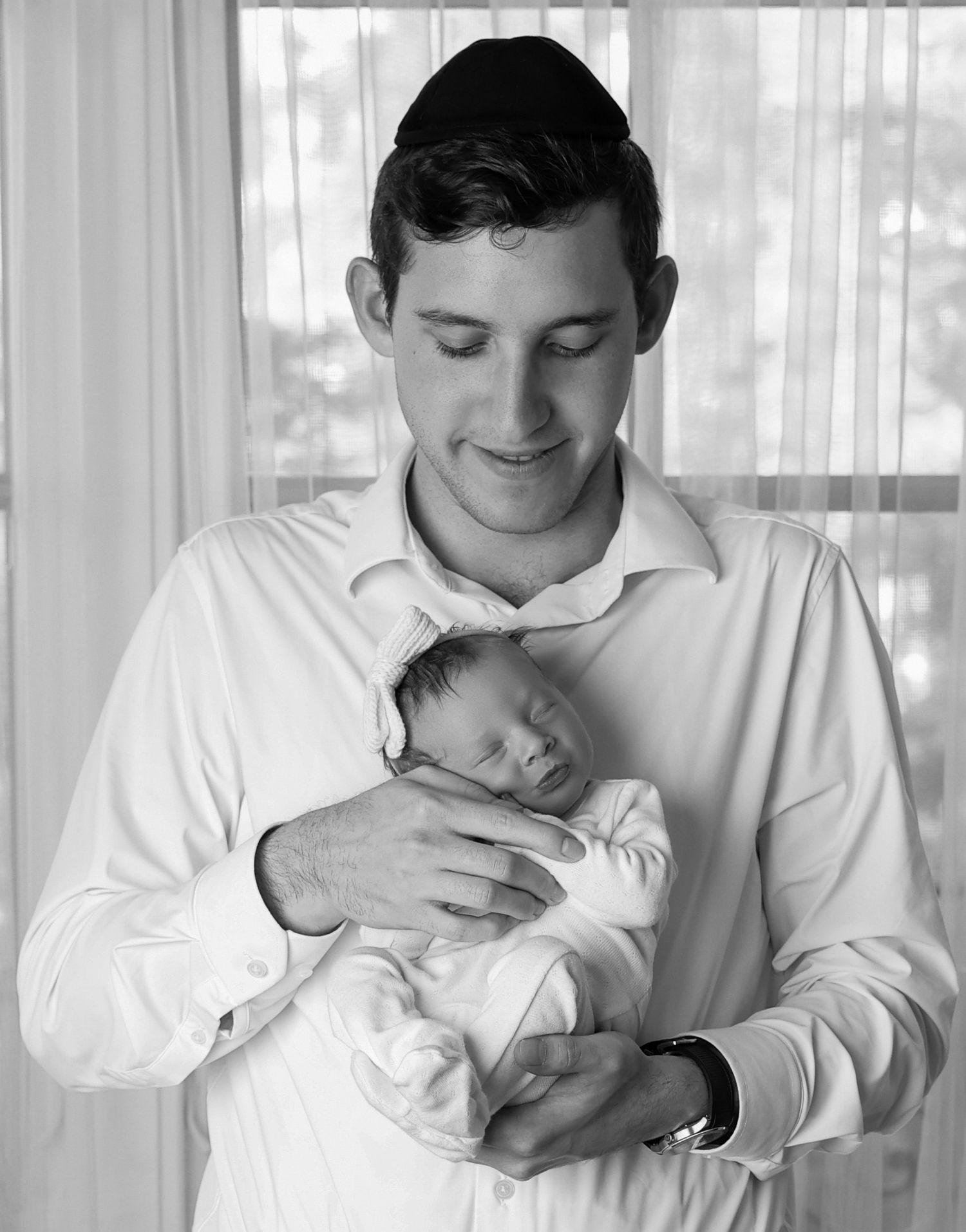 Tender black and white photo of a dad smiling down at his newborn baby girl in front of a window
