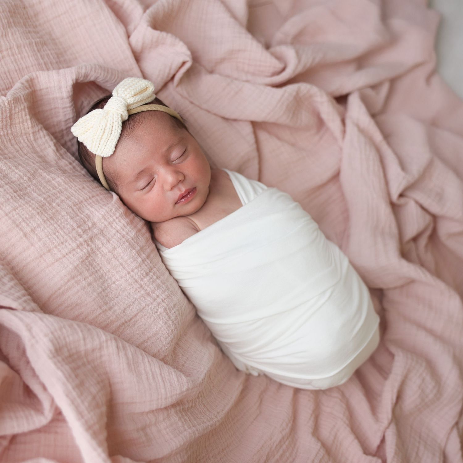 Classic baby girl newborn photography featuring a large white bow headband and tight swaddle wrap on a muslin pink background