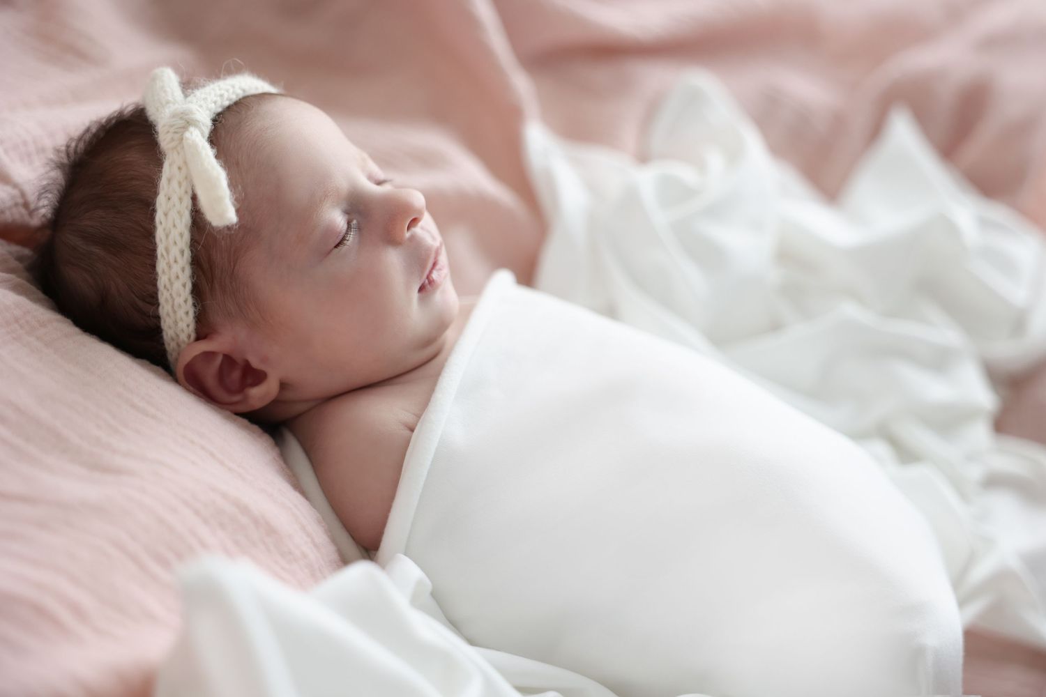 Newborn baby peacefully asleep on a soft pink blanket, wrapped in white fabric with a knitted bow.